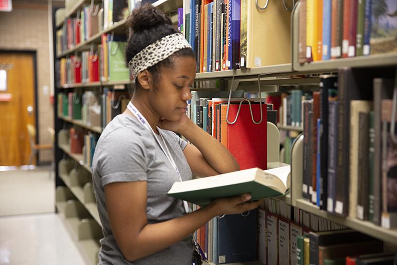 Photo of student in library reading book by shelves